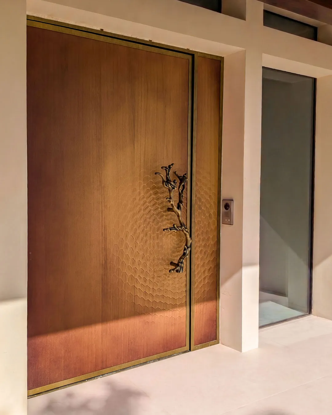 Porta d'ingresso su misura con apertura pivot, pannello fresato a nido d'ape, cornice OxyBrass e maniglia scultorea a ramo, realizzata da Metalbril
